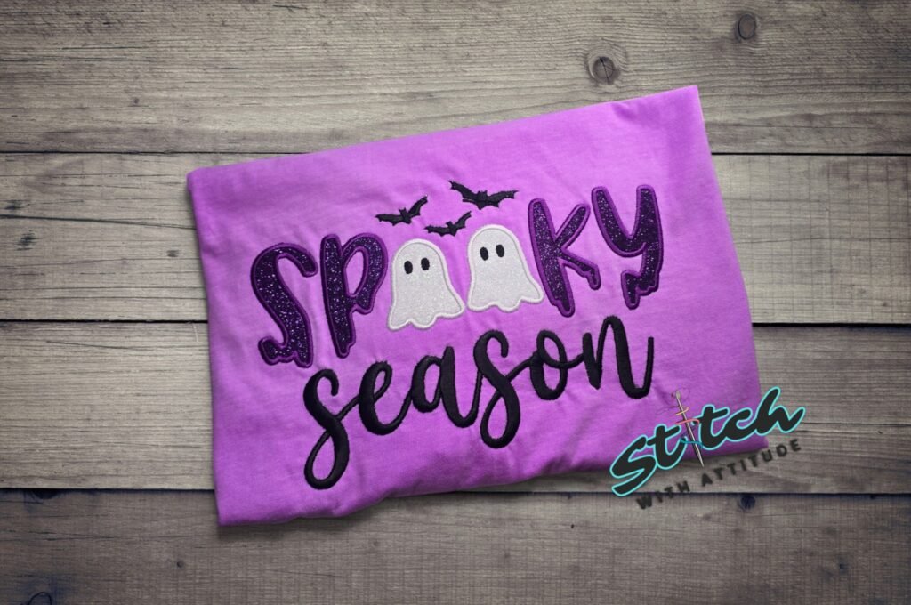 Spooky Season Glitter Ghost Applique Tee. SPOOKY in dark purple glitter, two little glitter ghosts standing in for the double O, and Season stitched in script underneath. Bats incl